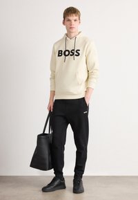 Jaunas vyras vilki smėlio spalvos BOSS džemperį, juodas sportines kelnes su BOSS logotipu, juodus sportinius batus, laiko didelę juodą rankinę prieš vientisą fono.