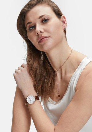 Runde Uhr mit weißem Zifferblatt, dünnen roségoldenen Akzenten und einem braunen Lederarmband. Das Modell trägt ein ärmelloses weißes Oberteil und geschichteten Schmuck.