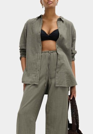 Vrouw draagt een losse linnen overhemd en broek in olijfgroen met opgerolde mouwen, zwarte bralette en houdt een donkergeweven tas vast.