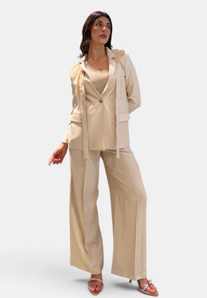 Donna con pantaloni beige a gamba larga, blazer abbinato con dettaglio a volant, top in maglia e tacchi aperti, in piedi davanti a uno sfondo chiaro e uniforme.