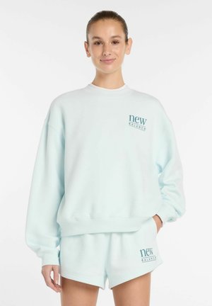 Jonge vrouw draagt een lichtblauwe New Balance trui en bijpassende korte broek, staat met één hand in haar zak en lacht tegen een witte achtergrond.