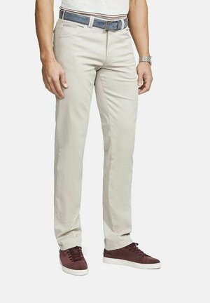 Mann trägt beige Chinos, blauen Gürtel, weißes Hemd, silberne Uhr und braune Sneakers, steht vor weißem Hintergrund.