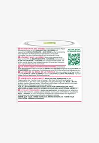 Kwadratowy biały pojemnik z metalową pokrywką, z kolorowym napisem i kodem QR. Etykiety w języku niemieckim i francuskim opisujące korzyści kremu.