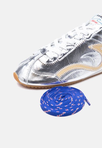Ασημί μεταλλικό αθλητικό παπούτσι με μπεζ αστέρι, λευκά κορδόνια και τυλιγμένο μπλε κορδόνι με κόκκινες και λευκές λεπτομέρειες σε λευκό φόντο.