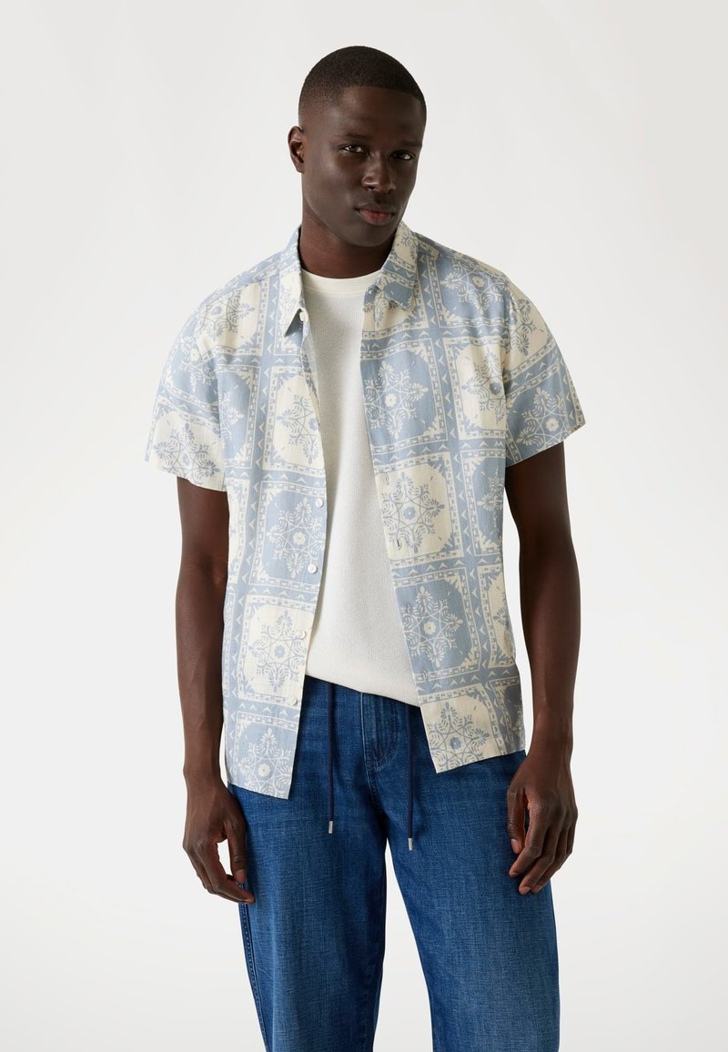 Camicia a maniche corte con bottoni, caratterizzata da un motivo floreale azzurro chiaro e crema, abbinata a pantaloni in denim, realizzata in tessuto traspirante.