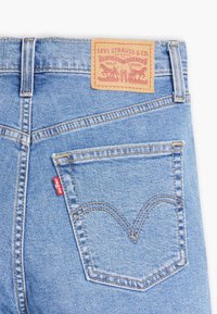Jeans de mezclilla azul claro con una etiqueta de parche de cuero en la cintura, que presentan una etiqueta roja en el bolsillo trasero y patrones de costura decorativa.