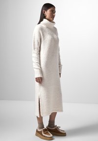 Abito maglione lungo, bianco, con texture, colletto alto a cappuccio, maniche lunghe e spacco laterale. Abbinato a scarpe marroni senza lacci e calze beige.