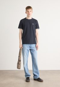 Tamsiai mėlynas medvilnės marškinėlis su baltais rausva apykaklės ir rankovių, šviesiai mėlynos džinsai, juodi sportiniai batai ir mažas rudas petinis krepšys.