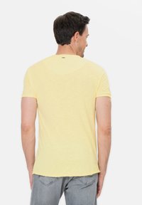Żółta koszulka z krótkim rękawem, okrągłym dekoltem i lekko teksturowanym materiałem. Z tyłu znajduje się subtelne logo nad szyją.