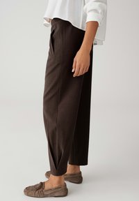 Braune, maßgeschneiderte Hose mit weitem Schnitt, versehen mit Falten und glatter Textur. Kombiniert mit grauen Mokassins.