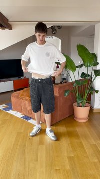 Jovem dentro de casa a levantar a camisa branca, vestindo calções pretos, meias brancas e ténis prateados, em pé sobre o chão de madeira perto de um sofá laranja e de uma grande planta em vaso.