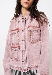 Pale roter Denimjacke mit zwei Brusttaschen, silbernen Knöpfen und grünem Akzent-Tag. Sie hat eine lässige Passform und eine ausgewaschene Textur.