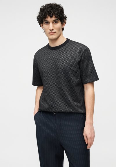 Άντρας με σγουρά μαλλιά, ντυμένος με μαύρο πουκάμισο με κοντά μανίκια και σχέδιο, και σκούρο παντελόνι με ρίγες, όρθιος με τα χέρια στις τσέπες.