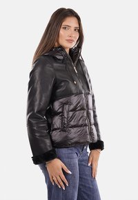 Giacca nera con cappuccio, con parte superiore in pelle e parte inferiore imbottita e trapuntata, dotata di polsini morbidi e accessori in metallo dorato. Texture liscia nel complesso.