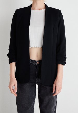 Person iført en sort blazer over en hvid croppet ribbet top og sort højtaljede jeans, stående mod en ensfarvet baggrund.