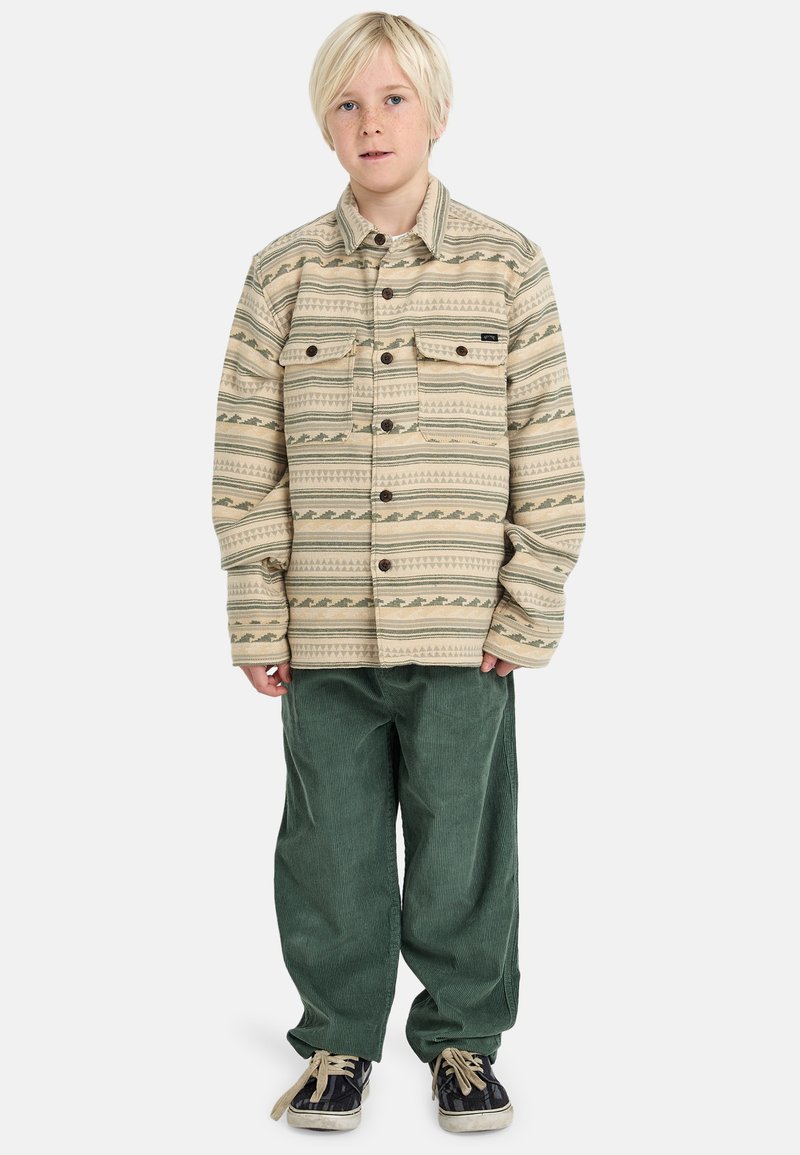 Giacca beige con motivo, dotata di due tasche frontali, pantaloni in corduroy verdi e scarpe da ginnastica nere. La giacca presenta un design geometrico e bottoni.