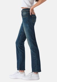 Pantalones vaqueros de mezclilla oscura con corte recto, cinco bolsillos y un ligero desgastado. Combinados con zapatillas blancas, resaltando la textura y el ajuste.