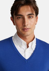 Blauer V-Ausschnitt-Pullover aus weichem Material, über einem weißen Hemd mit Kragen getragen. Verfügt über eine glatte Textur und eine klassische Passform.