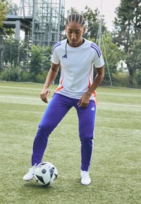 Adidas soccer outfit featuring a white and orange short-sleeve shirt, purple pants, and white shoes, with a black and white soccer ball on grass.