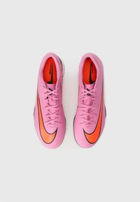 Scarpe da calcio rosa con superficie texture, caratterizzate da accenti swoosh Nike neri e arancioni. I lacci e le sole imbottite sono visibili.
