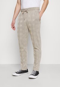 Beigefärgade rutiga byxor med elastisk midja och snörning. Tillverkade av textil med struktur och parat med svarta tygskor.