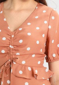 Pfirsichfarbenes Kleid mit weißen Punkten, V-Ausschnitt, gerafftem Vorderteil, kurzen Puffärmeln und einem fransigen Saum. Weicher, leichter Stoff.