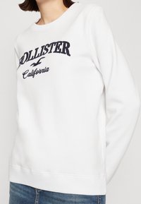 Vit sweatshirt med marinblå broderad text "Hollister California." Har rund halsringning och långa ärmar, tillverkad av ett mjukt, slätt material.