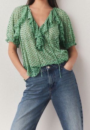 Woman wearing a sheer green ruffled blouse with puff sleeves and blue high-waisted jeans, hands tucked into front pockets.