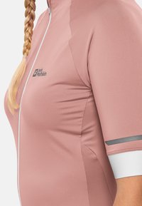 Haut de sport zippé à manches courtes rose, fabriqué en tissu lisse, avec un logo sur la poitrine et des accents réfléchissants sur les manches.