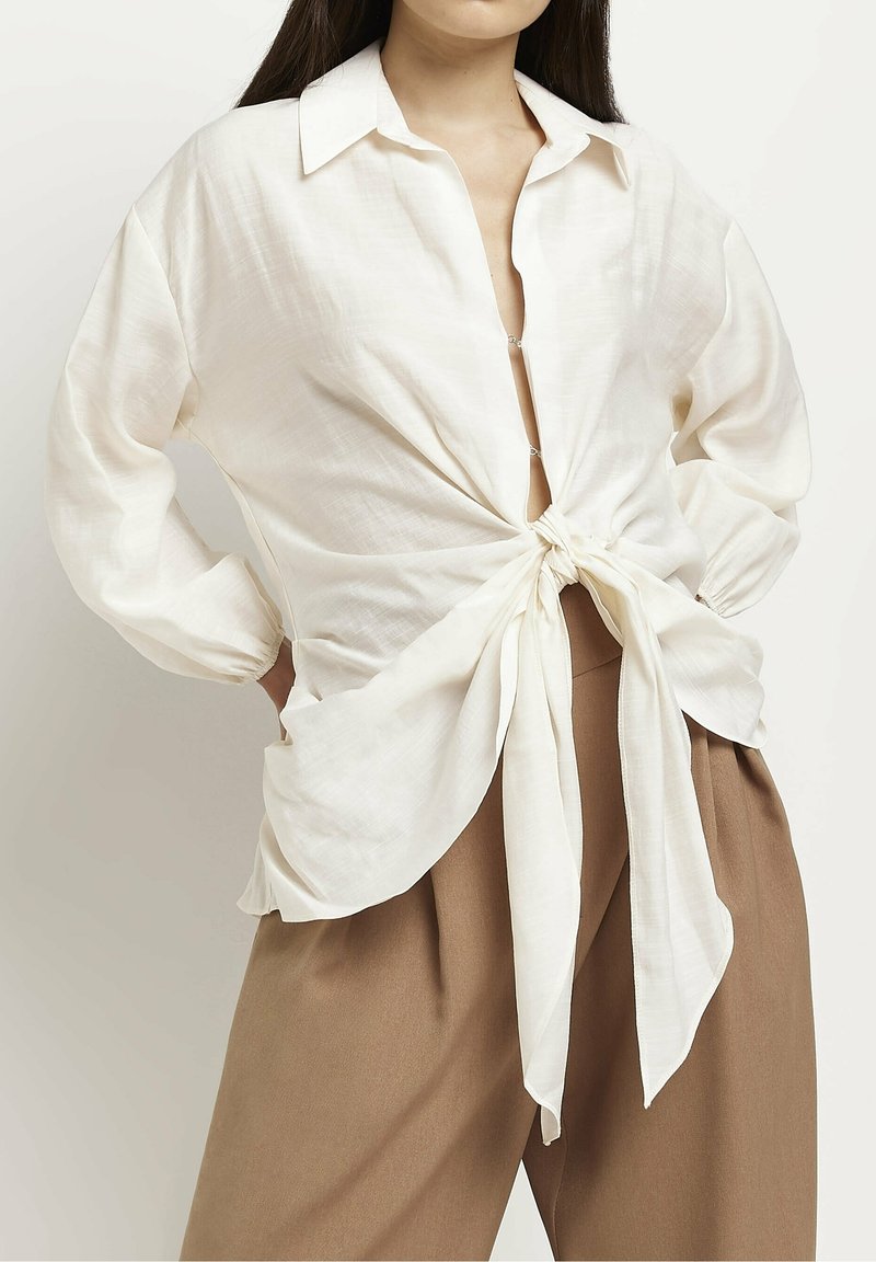 White sheer blouse with a knotted front detail, collared neckline, and long sleeves. Paired with light brown high-waisted trousers.