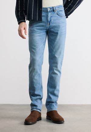 Man wearing light blue slim-fit jeans, brown suede ankle boots, and a dark jacket with white vertical stripes, standing on a gray floor.