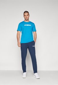Camiseta azul de manga corta con el logo "HUMMEL", combinada con pantalones de chándal marinos con acentos de logo, llevada con zapatillas deportivas blancas.