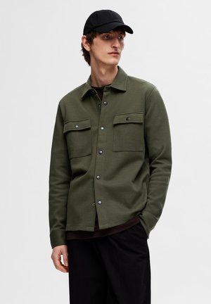 Olive green button-up shirt with two chest pockets, made of soft fabric, features snap buttons and a rounded hem, layered over a dark shirt.