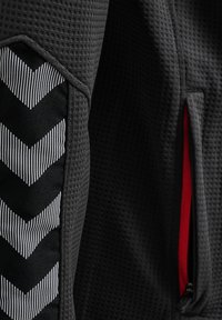 Nahaufnahme eines dunkel strukturierten Stoffes mit schwarz-weißem Chevron-Design und einer Reißverschlusstasche, die das rote Innenfutter zeigt.