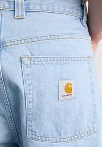 Lysblå denimshorts med en synlig baglomme, prydet med et hvidt mærke-logo med gule accenter. Syede detaljer i en kontrastfarvet tråd.