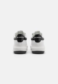 Zapatillas blancas de cuero con acentos de charol negro, punta redonda, collar acolchado y suela de goma texturizada. Marca visible en el talón.