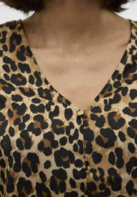 Top beige con design a stampa leopardata con macchie nere e marroni, scollatura a V e chiusura centrale con bottoni. Tessuto testurizzato.