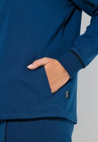 Top deportivo azul de algodón con un bolsillo lateral, puños acanalados y una pequeña etiqueta con el logo en el dobladillo. Textura suave con un ajuste relajado.