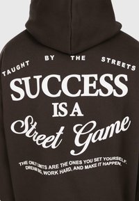 Hoodie noir avec texte blanc : "Enseigné par la rue. Le succès est un jeu de rue. Les seules limites sont celles que vous vous imposez. Rêvez grand, travaillez dur."