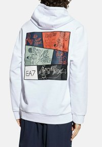 Personne portant un sweat à capuche blanc avec des panneaux de bande dessinée sportifs colorés dans le dos, présentant des mascottes de basket-ball, de football, de tennis et de golf.