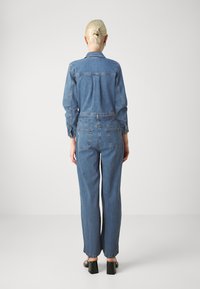 Denim overáll, világoskék, hosszú ujjak, gombos elöl, rugalmas derékpánt, bő szárú nadrág, két hátsó zseb, fekete magassarkú szandállal viselve.