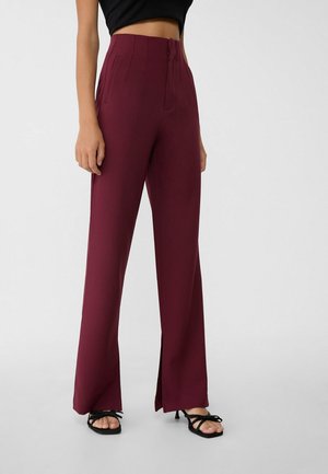Femme portant un pantalon bordeaux taille haute à jambes larges avec des fentes à l'avant et des sandales à talons noires à brides, sur un fond uni.
