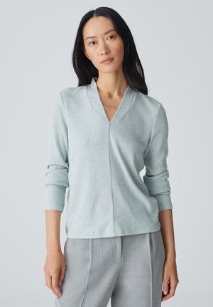 Maglione a coste blu chiaro con scollo a V e maniche lunghe. Texture morbida, design pulito, indossato con pantaloni grigi chiari.