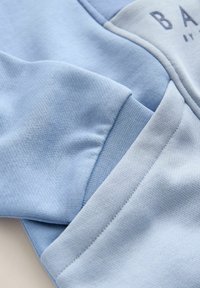 Material albastru deschis, prim-plan, arătând detalii texturate de tricot și cusături ale unui articol vestimentar pliat, cu text parțial imprimat în partea superioară dreaptă.