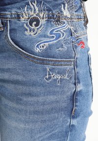 Jeans de mezclilla de color azul con diseños de dragones bordados. Incluye parches y detalle de logo en hilo blanco en el bolsillo izquierdo.