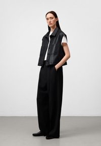 Gilet in pelle nera con colletto, maniche corte e due tasche frontali, indossato sopra una maglietta bianca e abbinato a pantaloni neri larghi.
