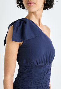 Tums zila vienas pleca kleita ar lielu loku detaļu, teksturētu drapējumu uz ķermeņa un mīkstu audumu. Tuvplāns uz augšējo daļu.