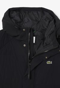 Chaqueta acolchada negra con cuello alto, cierre de cremallera y botones a presión. Presenta un logo de Lacoste y un forro interior liso y acolchado.