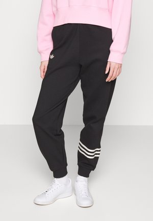 Person wearing black Adidas sweatpants with three white stripes near the cuff and white sneakers, paired with a pink sweatshirt.