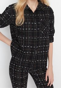 Camicia nera con bottoni e motivo a quadri, con accenti bianchi e rossi. La camicia ha un colletto, maniche arrotolate e una tasca sul petto.